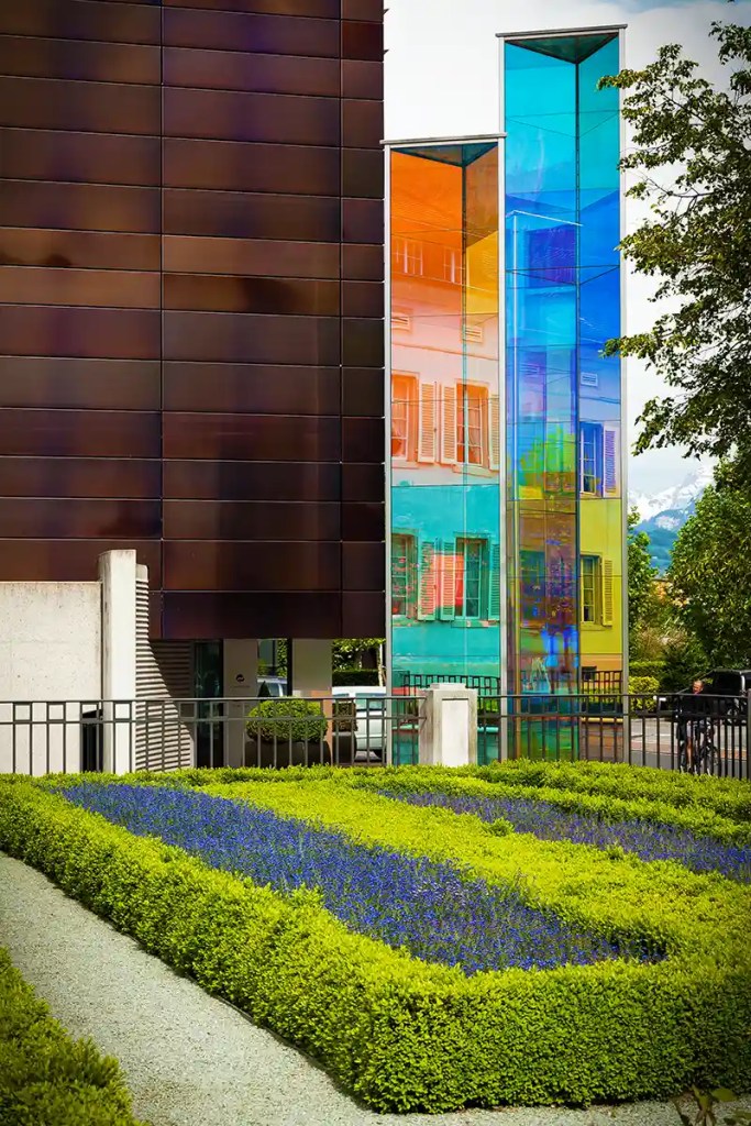 Modern building with dark metal panels beside tall glass columns tinted in blue, orange, and green that reflect the surrounding street. In front of the structure, neatly trimmed hedges and rows of purple flowers form a geometric garden along a gravel path.