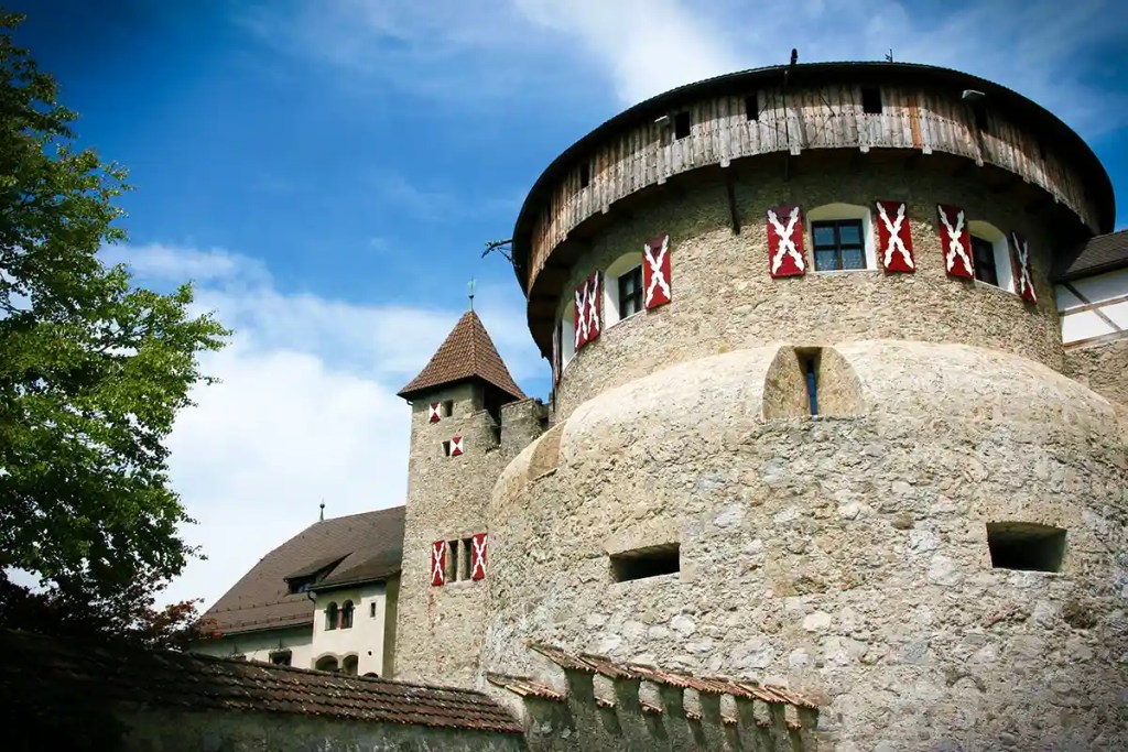 Round stone tower with small windows and red shutters featuring white cross patterns, attached to a historic fortress style building. The thick textured walls and narrow openings suggest defensive architecture, while a smaller tower with a pointed roof rises behind it against a partly cloudy sky.
