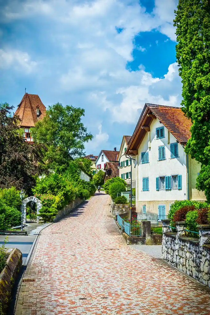 Charming cobblestone street lined with traditional houses featuring sloped roofs and pastel shutters, surrounded by lush greenery and trees. A small stone pathway curves gently uphill toward more homes and a tower in the distance, under a sky filled with soft clouds and patches of blue.