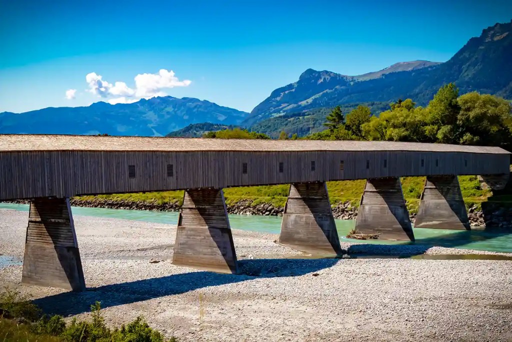Long wooden covered bridge supported by concrete pillars stretches across a shallow turquoise river with a wide gravel riverbed. Forested hills and blue mountains rise in the background beneath a bright clear sky.
