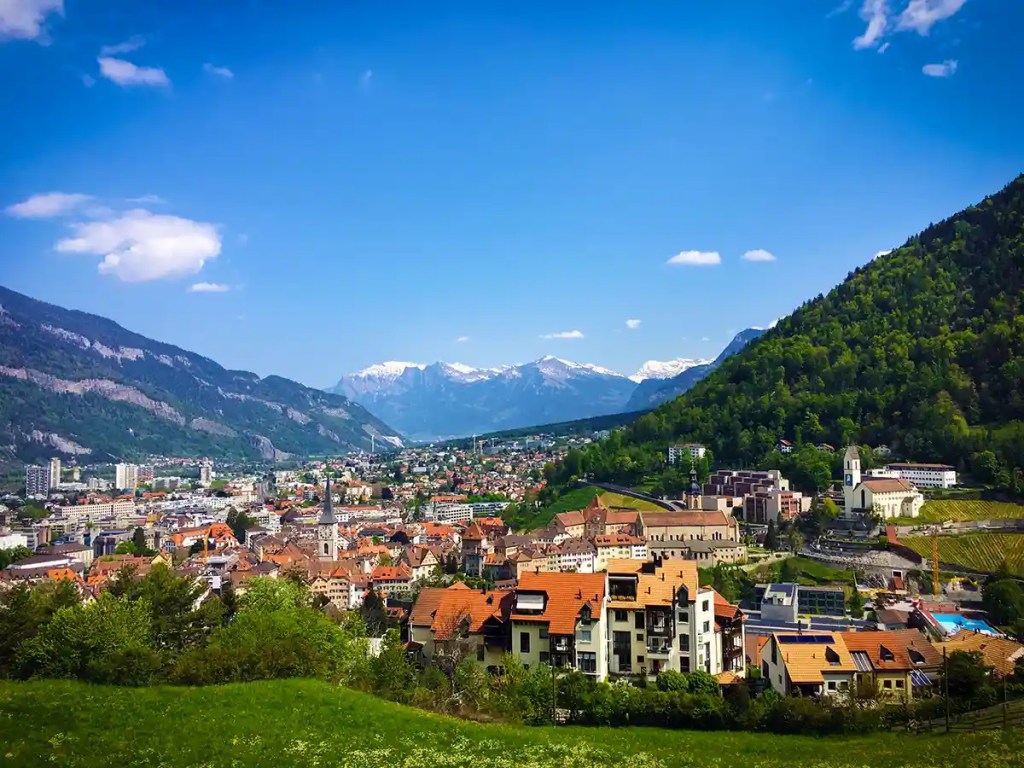 Panoramic view of a town with red tiled roofs and church spires spread across a valley between green forested hills. Snow capped mountains rise in the distance under a clear blue sky with a few small clouds.