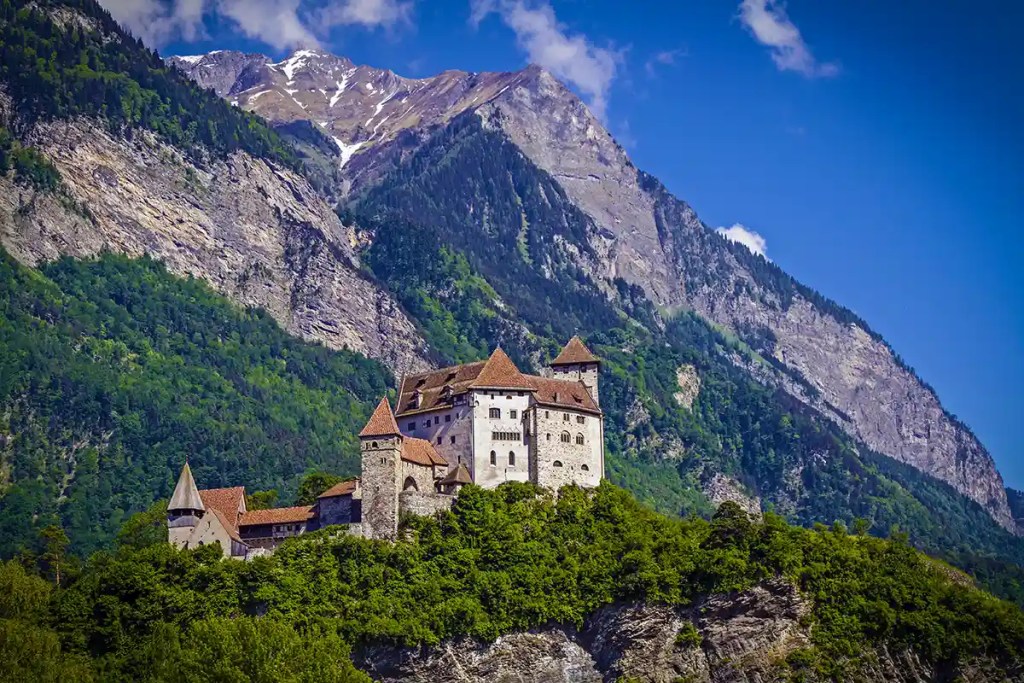 Medieval stone castle with red tiled roofs and round towers perched on a wooded hill. Towering rocky mountains rise steeply behind the fortress under a deep blue sky with scattered clouds.