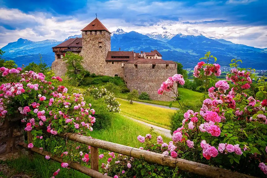 Stone hilltop castle with a square tower and red tiled roofs overlooking green fields and a winding road. Pink rose bushes and a wooden fence frame the foreground while snow capped mountains rise in the distance beneath dramatic clouds.