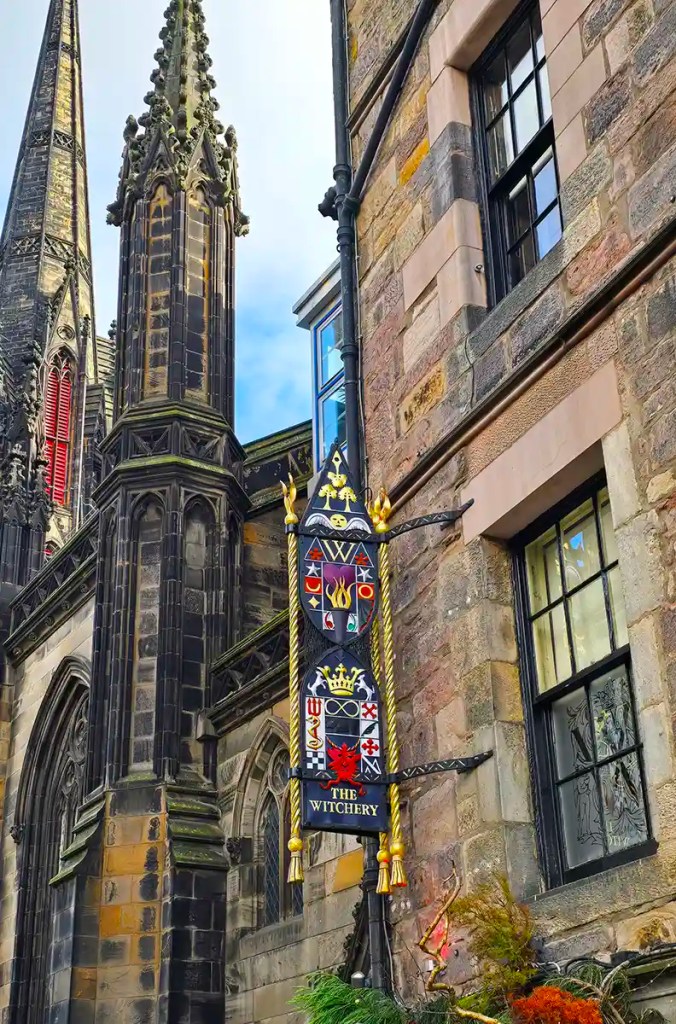 Ornate hanging sign reading "THE WITCHERY" with decorative symbols and crests, mounted on a stone building beside a gothic church with tall spires. Detailed stonework and stained glass windows frame the narrow street scene, highlighting historic architecture.