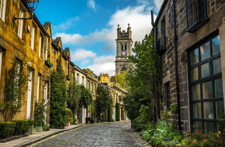 Charming cobblestone street lined with historic stone houses covered in greenery, leading toward a tall clock tower in the distance. Warm sunlight highlights the textured buildings and plants along the quiet, curved road under a bright blue sky with scattered clouds.