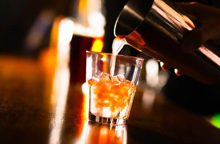 Close up of a drink being poured from a metal cocktail shaker into a short glass filled with ice on a dimly lit bar counter. Warm orange and amber lighting reflects off the glass and surface, creating a moody nightlife atmosphere.