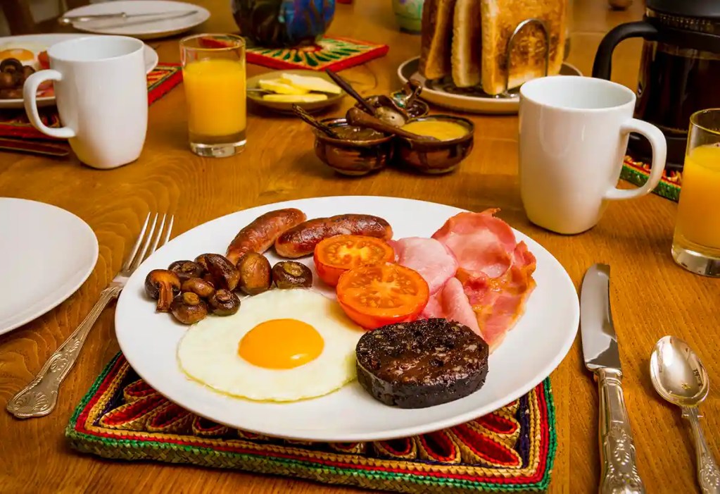 Traditional Scottish breakfast with fried egg, sausage, bacon, black pudding, mushrooms, and grilled tomatoes served on a white plate.