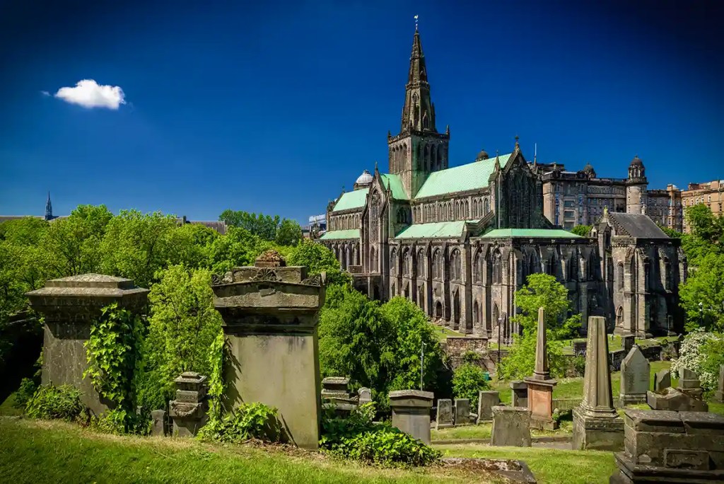 View of Glasgow Cathedral and surrounding gravestones from the historic Glasgow Necropolis cemetery on a sunny day.