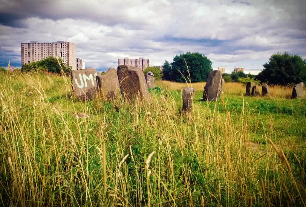 Weathered gravestones and tall grass at the historic Sighthill Cemetery in Glasgow with apartment buildings in the background.