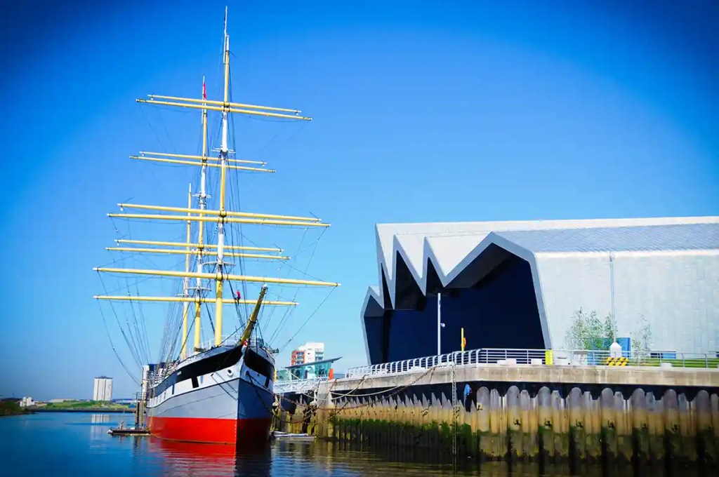 Tall ship docked beside the Riverside Museum in Glasgow along the River Clyde on a clear sunny day.