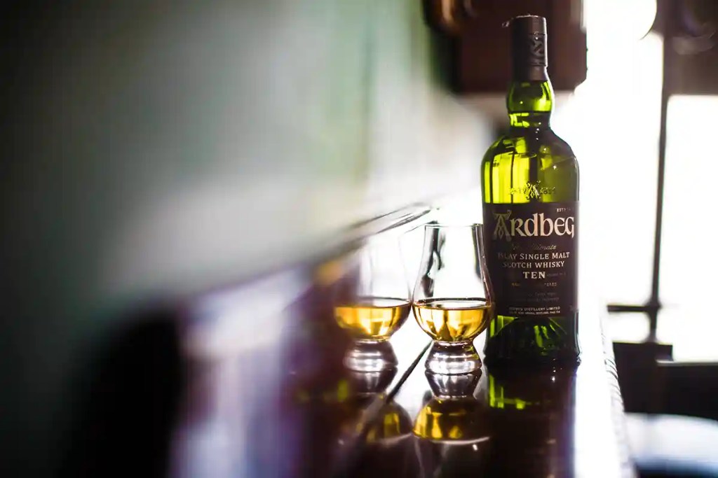 A green bottle labeled "Ardbeg Ten Islay Single Malt Scotch Whisky" stands beside two glasses of amber whisky on a reflective table. Bright window light creates deep shadows and reflections across the tasting room.