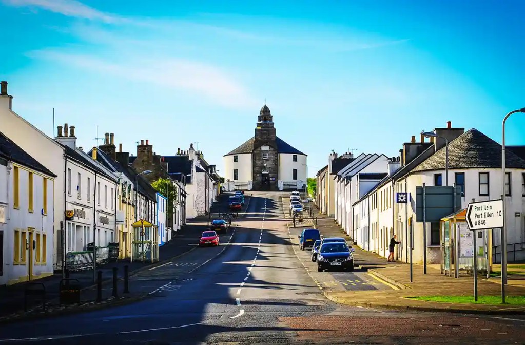 A quiet town street climbs uphill between rows of white buildings toward a round stone and white building at the crest. A road sign points right with the text "Port lein Port Ellen 10 A846" under a bright blue sky.