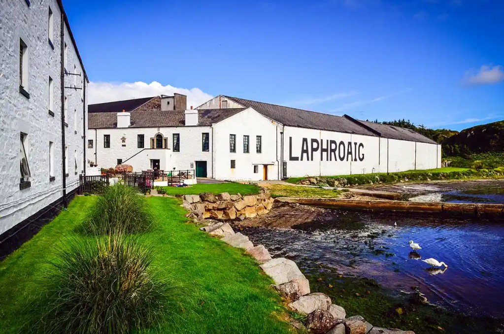 White distillery buildings marked "LAPHROAIG" sit beside a small inlet under a bright blue sky. Green grass rocky shoreline and two white birds on the water frame the coastal scene.