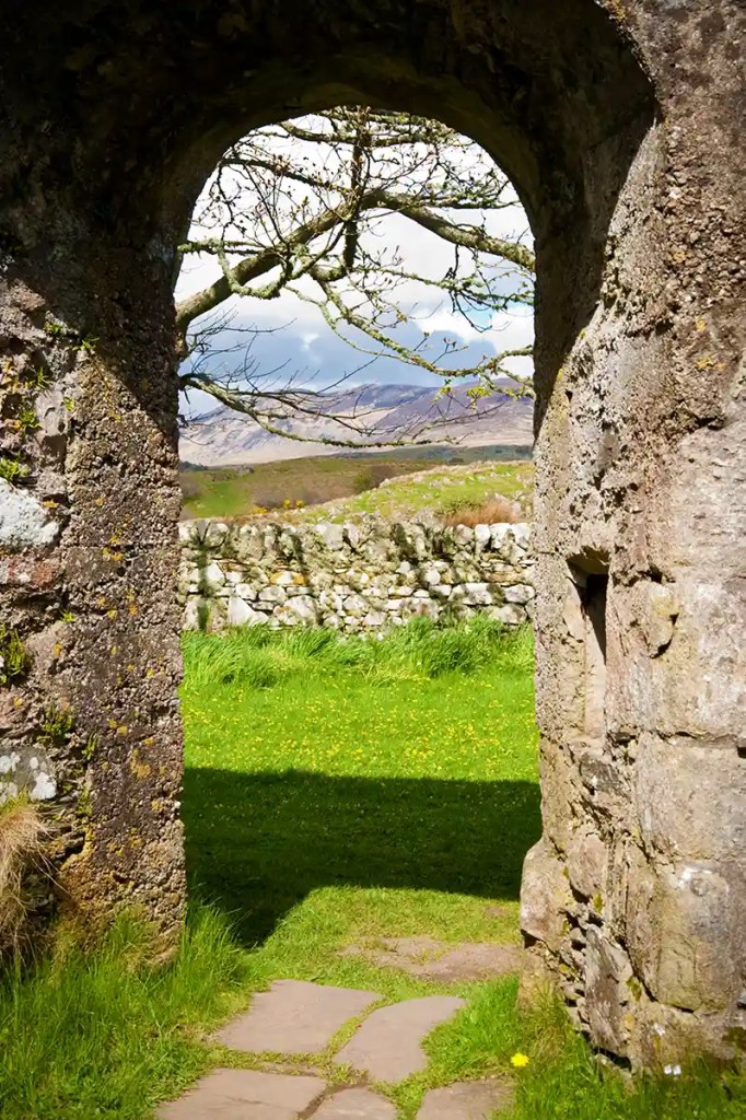 An old stone arch frames a view of bright green grass and a low stone wall. Bare tree branches stretch across the opening with hills and cloudy sky beyond.