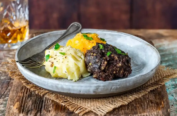 A rustic plate holds a dark crumbly savory portion beside creamy mashed potatoes and golden mashed root vegetables. Chopped parsley and black pepper garnish the meal with a glass of amber whisky in the background.