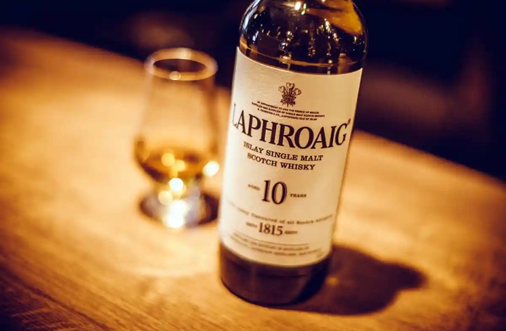 A bottle labeled "LAPHROAIG Islay Single Malt Scotch Whisky 10 years 1815" sits on a wooden table in warm low light. A whisky glass glows softly in the background with the bottle in sharp focus.