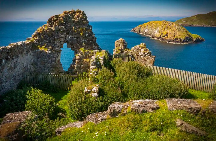 Weathered stone ruins with an arched opening stand above a vivid blue sea with a small rocky island just offshore. Green grass and dense plants surround the ruins with a wooden fence tracing the cliff edge.