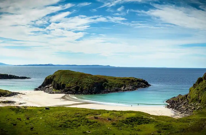 A white sand beach curves around clear turquoise water between steep green cliffs and a grass covered rocky island. Tiny figures walk near the shoreline while grazing animals dot the meadow beneath a bright cloud streaked sky.