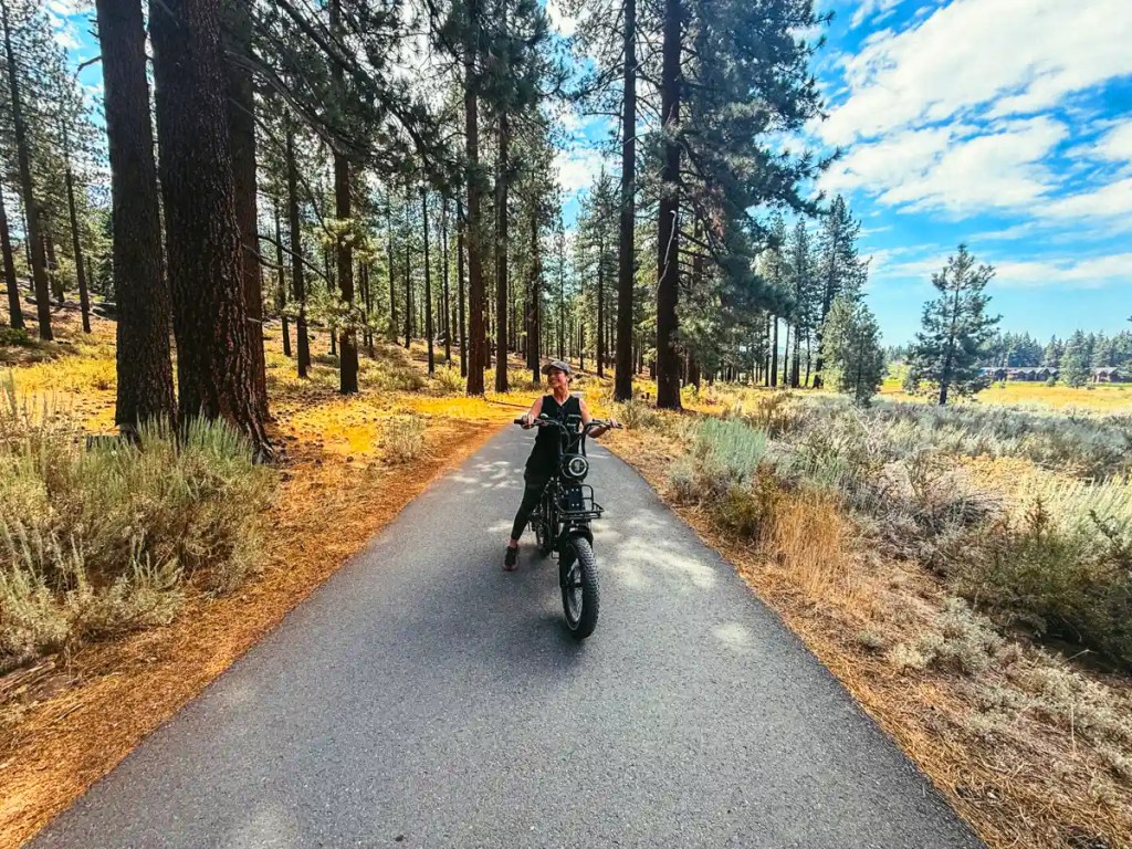 A woman stands with a black electric bike on a paved trail surrounded by tall pine trees and golden grasses. Bright blue sky and scattered clouds frame the sunny forest setting.