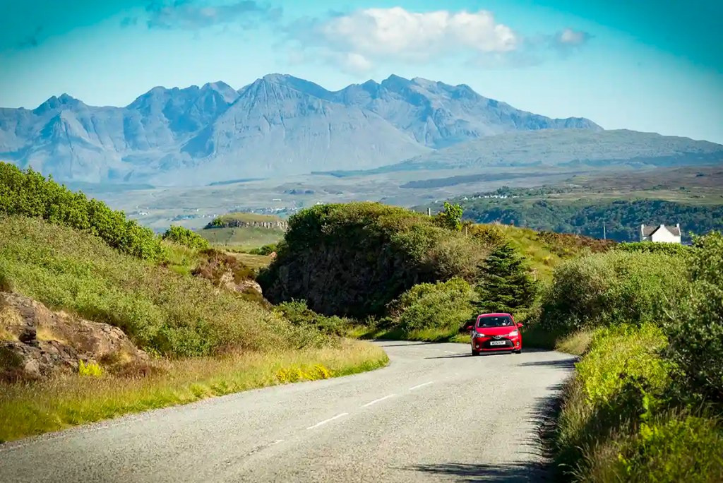 Red car driving along a winding rural road through green countryside, with dramatic rocky mountains rising in the distance.