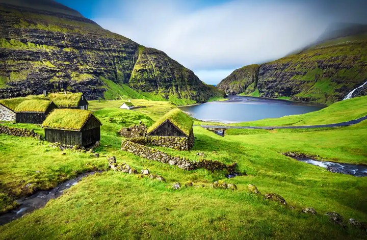 Traditional turf roofed cottages and stone walls scattered across a lush green valley in the Faroe Islands. A winding stream and narrow road lead toward a tranquil lake surrounded by steep rocky mountains, with a waterfall cascading down the hillside beneath low clouds.