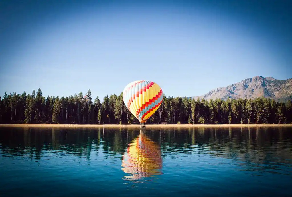 A colorful hot air balloon with red, yellow, and blue stripes floats just above a calm lake, casting a vibrant reflection on the water. Dense pine forests and rugged mountains frame the scene beneath a clear blue sky.