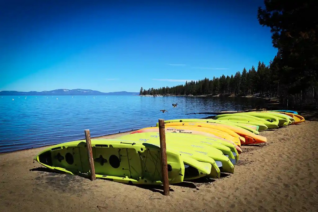 Bright green and yellow kayaks line a sandy beach beside a calm blue lake. Pine forests stretch along the shoreline, with distant mountains and a clear blue sky in the background.