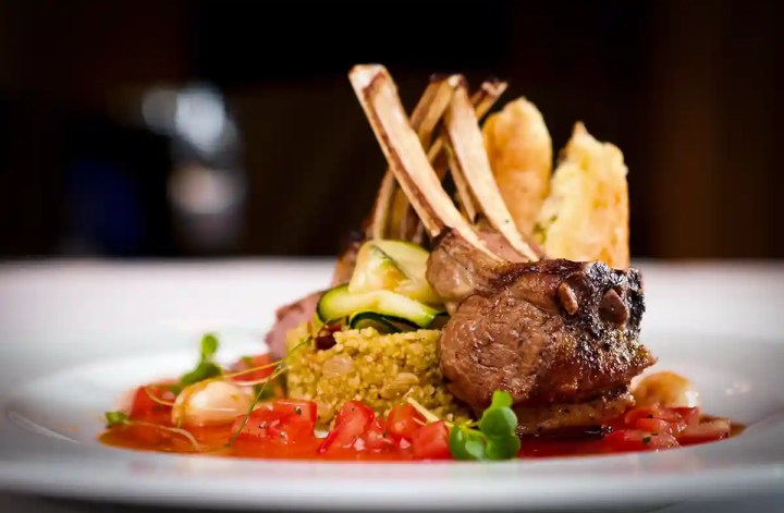Grilled lamb chops arranged over couscous with ribbons of zucchini and roasted vegetables. Diced tomatoes and fresh greens add color around the plated entrée.